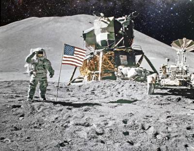 Image of astronaut on moon next to an American flag with a space craft in the background.  