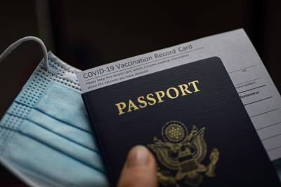 Image of a hand holding a passport, a face mask, and a COVID-19 vaccine record card. 
