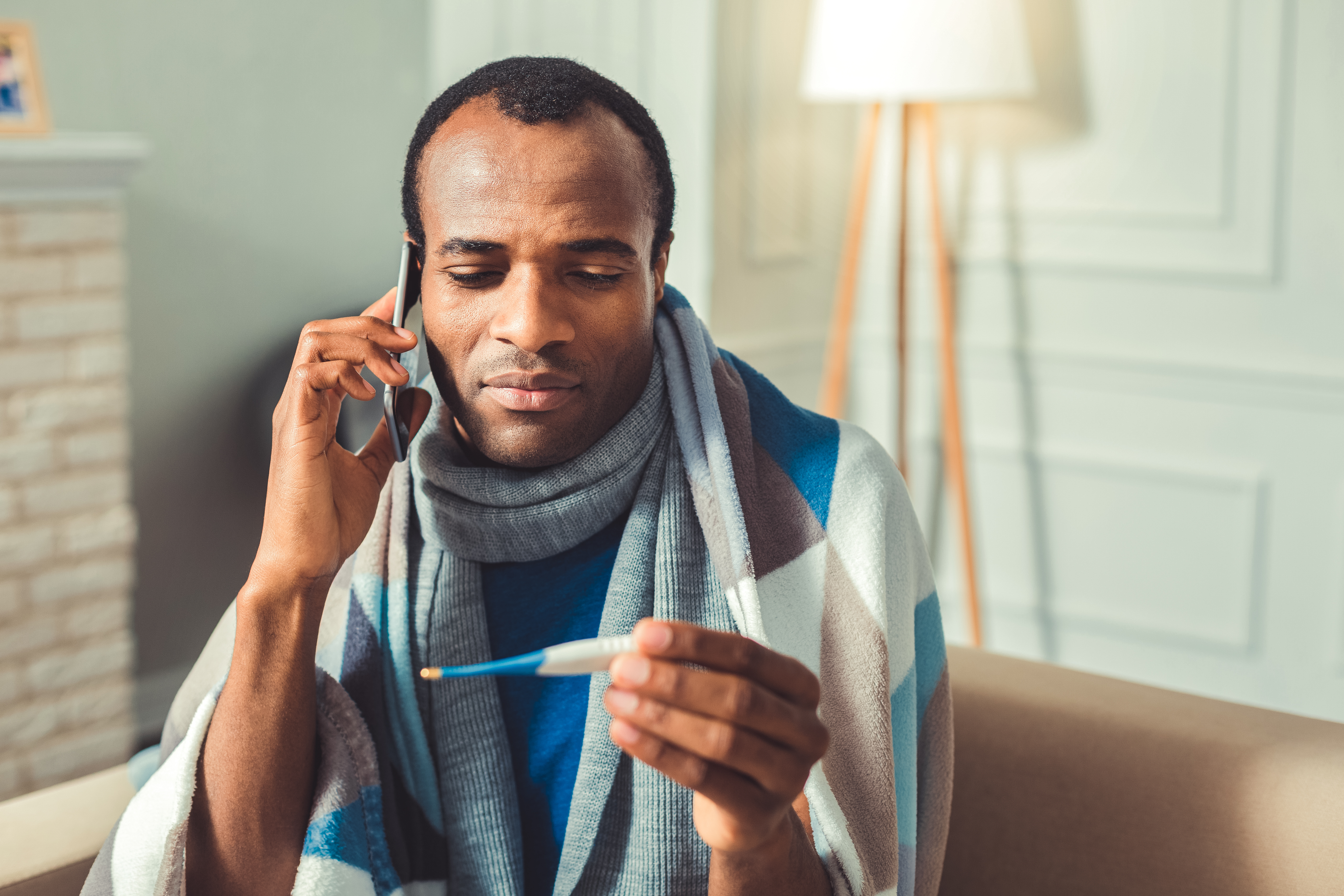 millennial man at home sick with scarf and thermometer talking on the phone