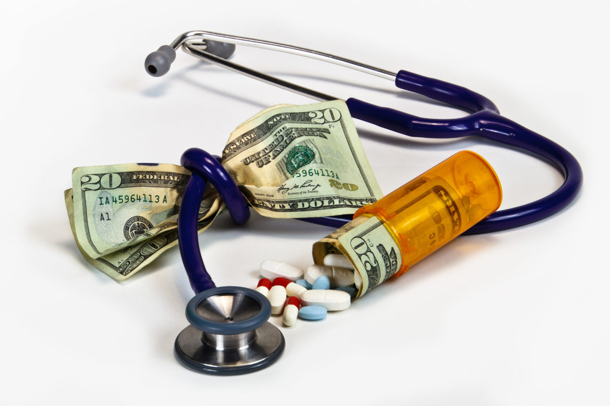 A stethoscope tied around a pile of cash, with a pill bottle nearby. The pill bottle has cash and pills inside.