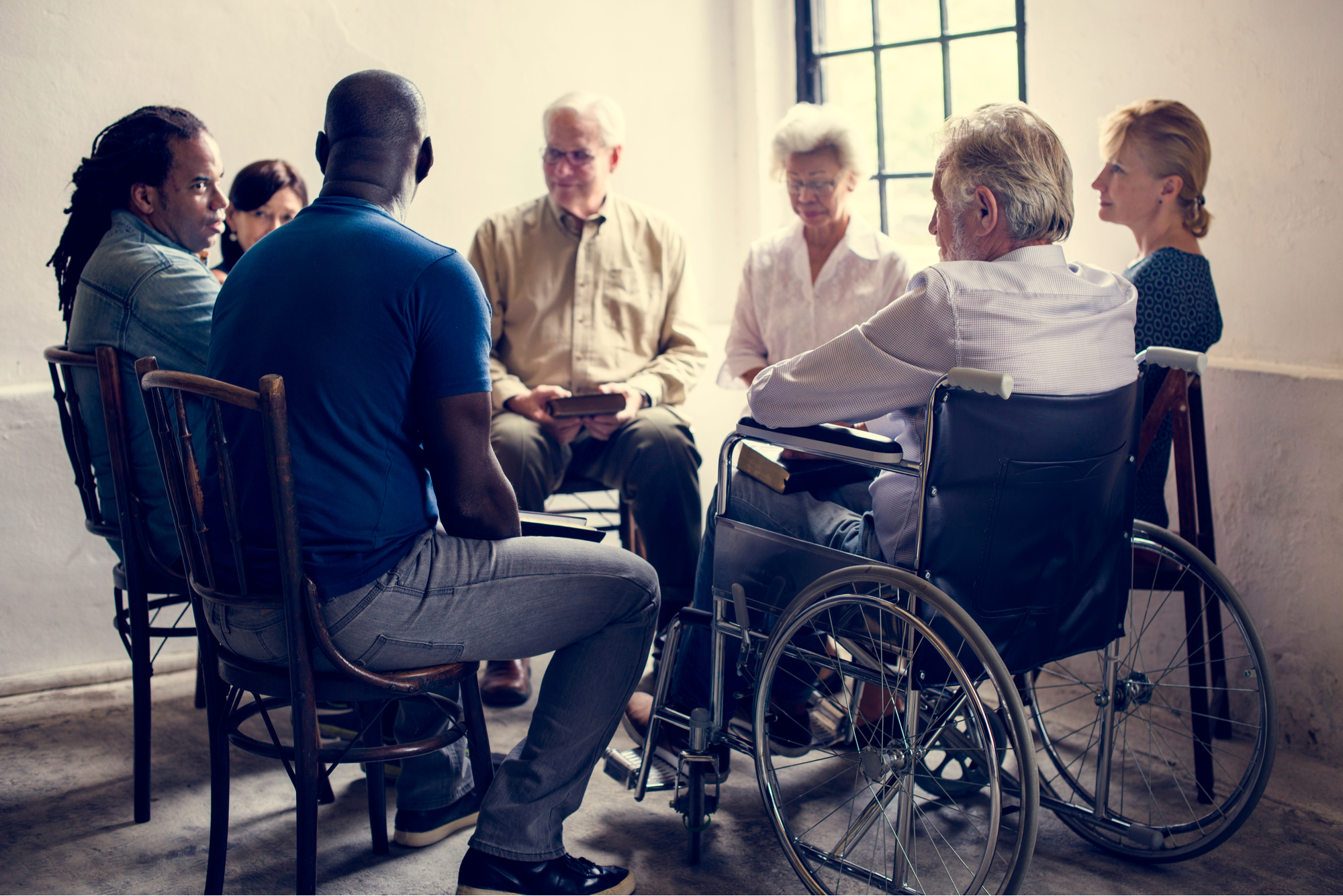 Group of people of various ages and ethnicities sitting in a circle talking. At least one of the people is in a wheelchair.