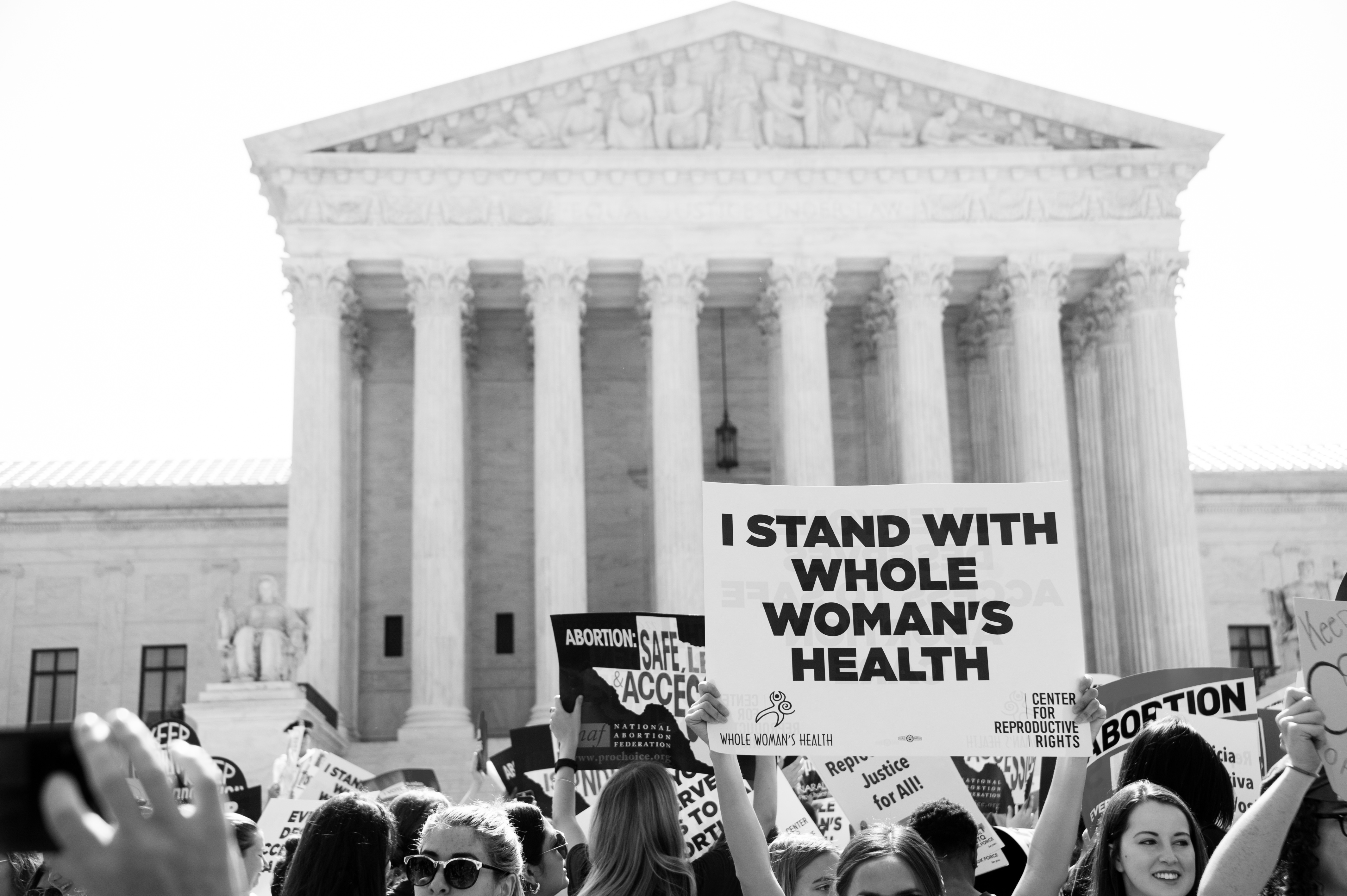 Black and white photograph of the front of the Supreme Court. Pro-abortion protestors stand holding signs, one of which reads "I stand with Whole Woman's Health"