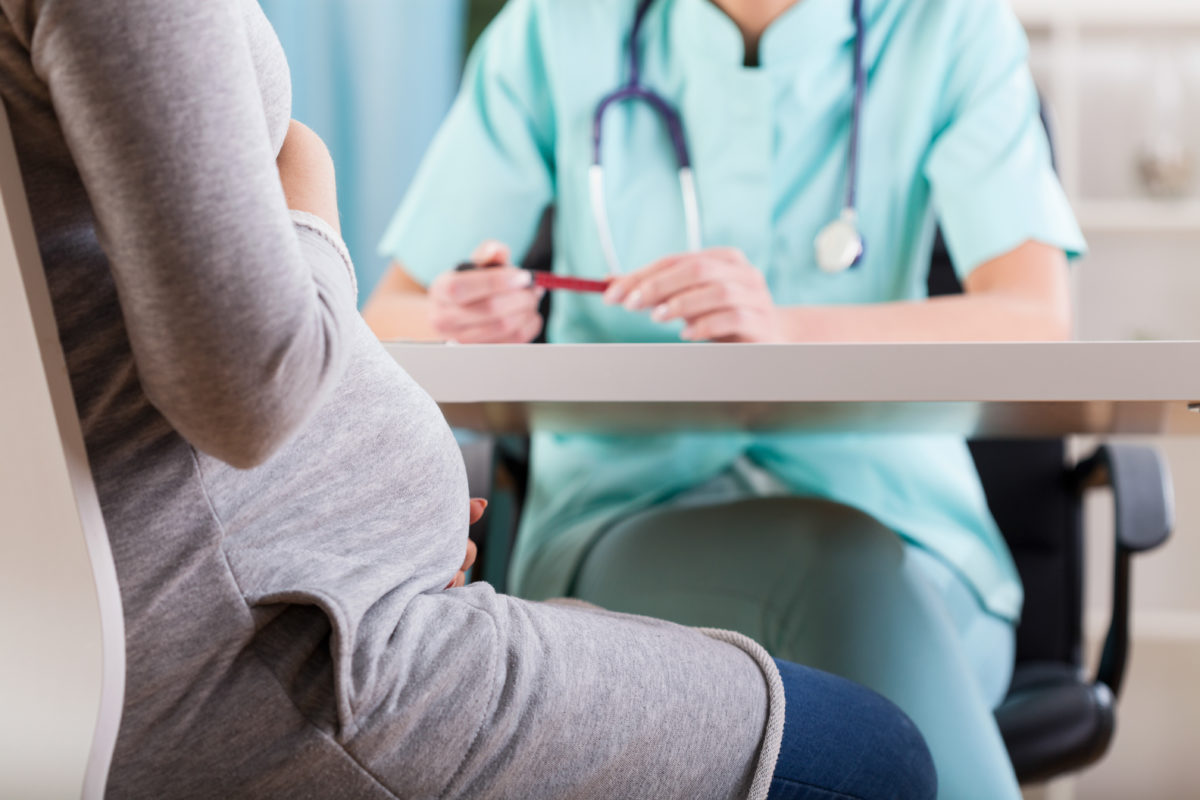 Pregnant woman sitting across desk from doctor wearing scrubs and holding a pen