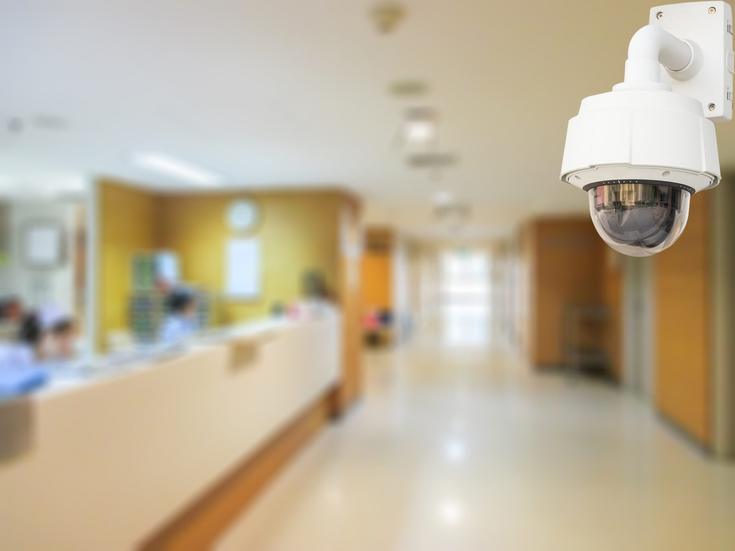 A surveillance camera in focus in the foreground of hospital hallway.