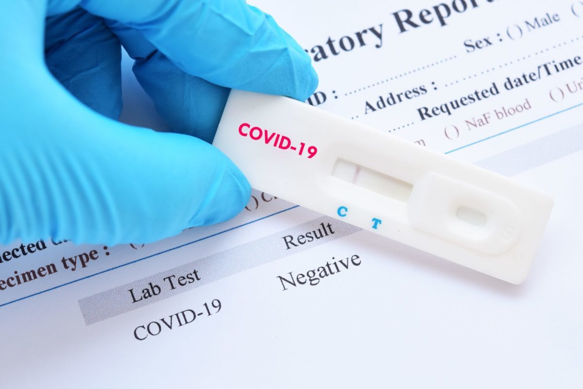 Gloved hand holding medical rapid test labeled COVID-19 over sheet of paper listing the test result as negative.