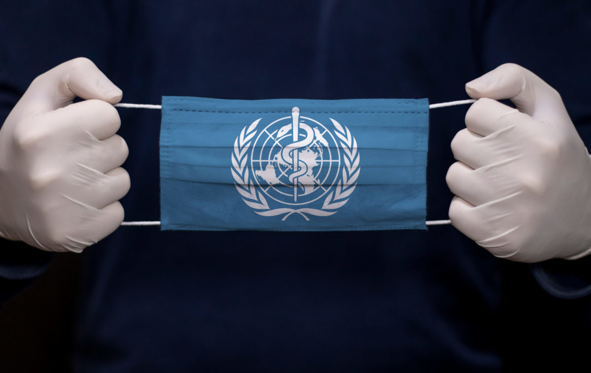 Gloved hands hold medical face mask with WHO (World Health Organization) flag.