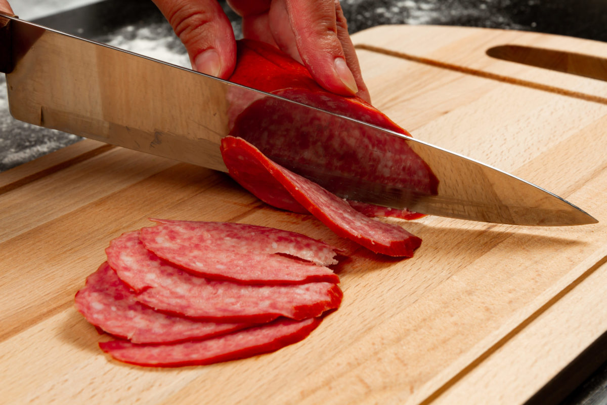 person cuts salami sausage on a wooden cutting board.