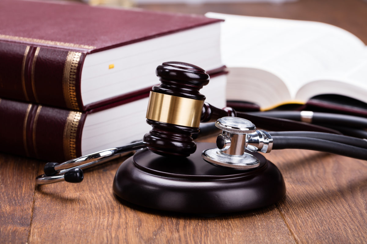 Brown Gavel With Medical Stethoscope Near Book At Wooden Desk In Courtroom.