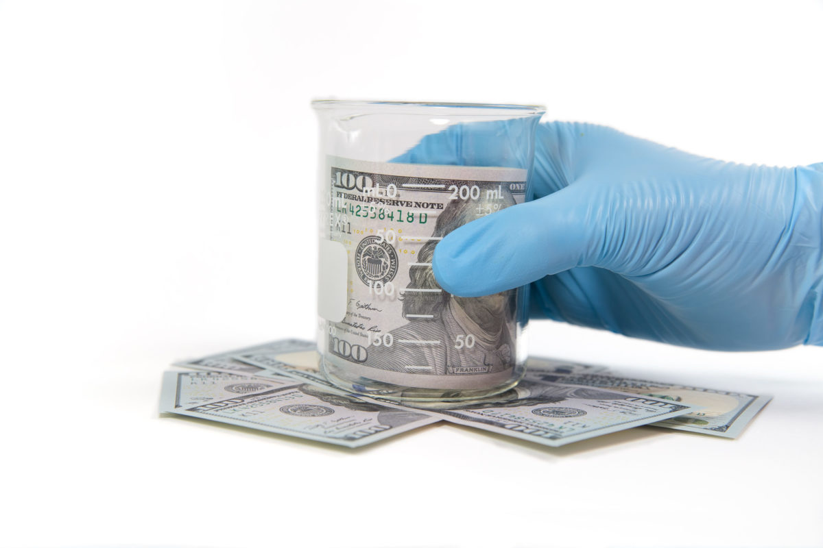 Gloved hand grabs beaker with rolled currency.