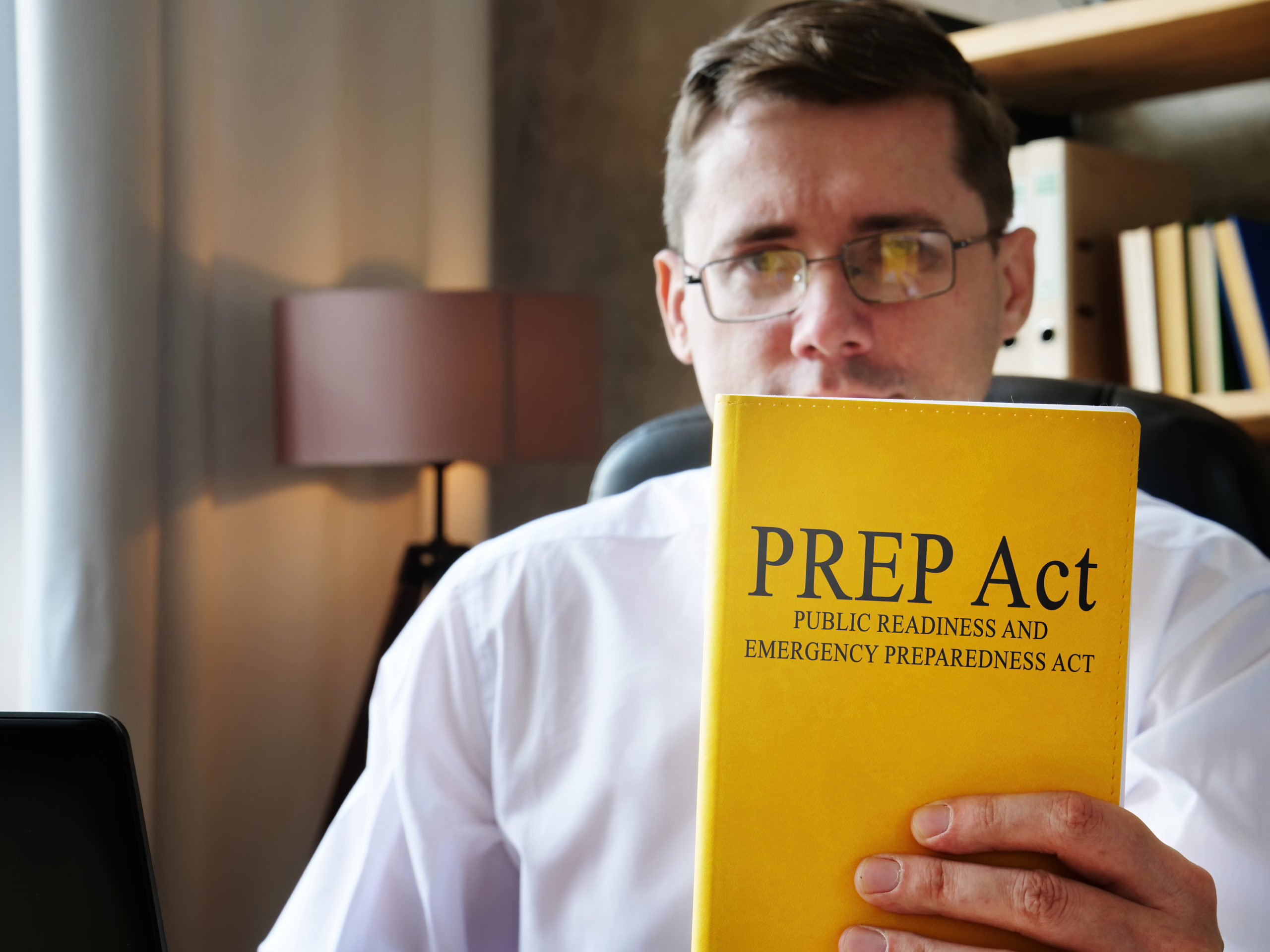 Bill of Health -man in glasses sitting in office holds yellow book labeled "PREP Act," PREP act pandemics, nursing home lawsuits