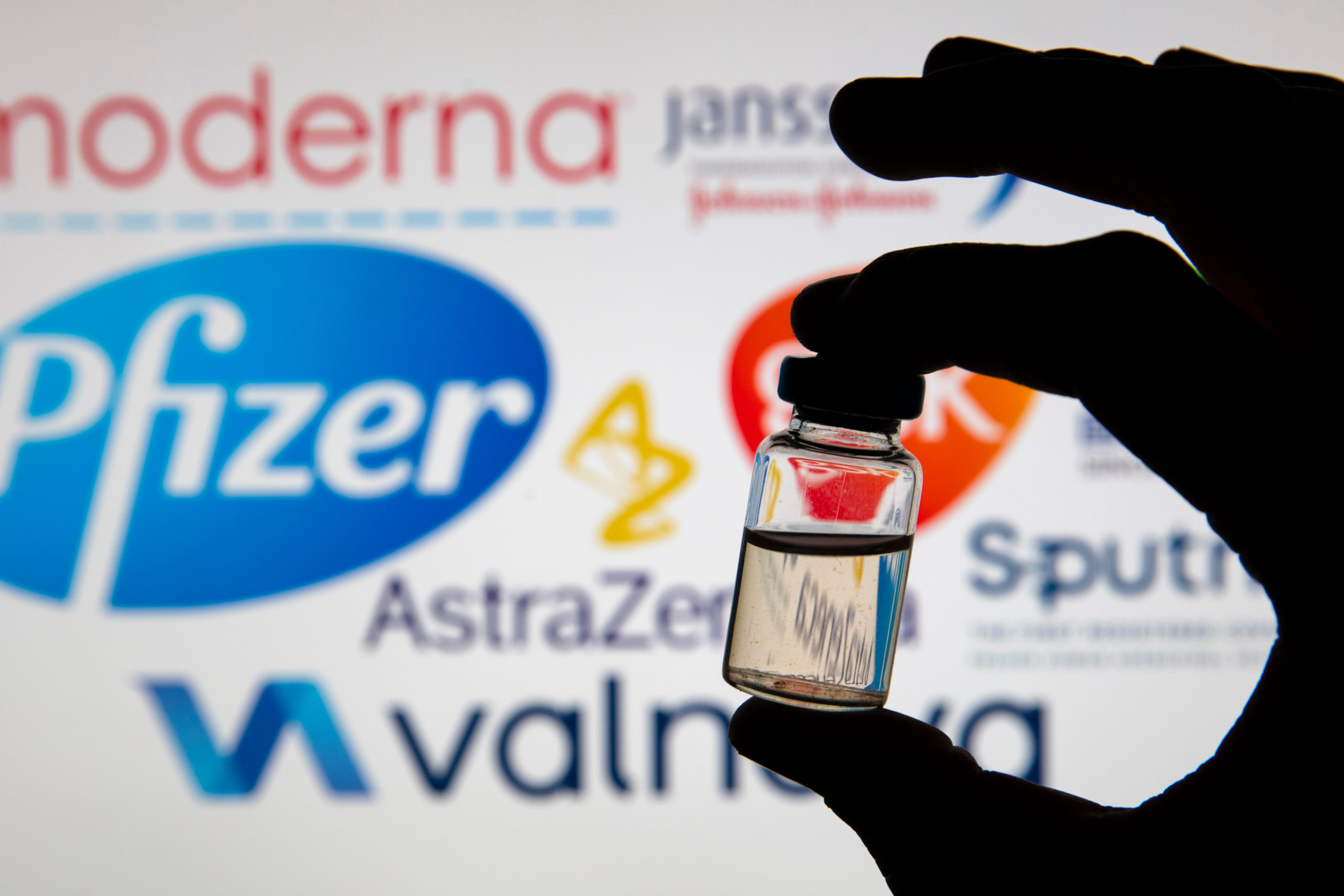 Bill of Health - silhouette of COVID-19 vaccine vile held in front of company logos, cooperation and antitrust in vaccine production