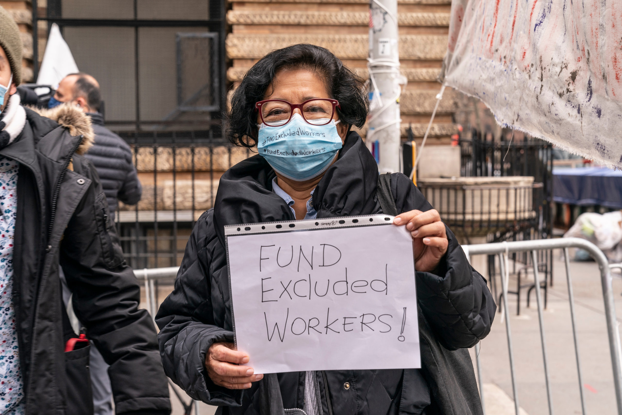 Bill of Health - Latina woman wearing mask holds protest sign saying "Fund undocumented workers," covid-19 pandemic undocumented workers