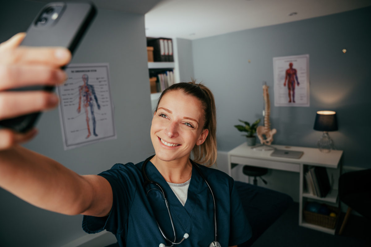 Caucasian female nurse standing in doctors office taking selfie on cellular device.