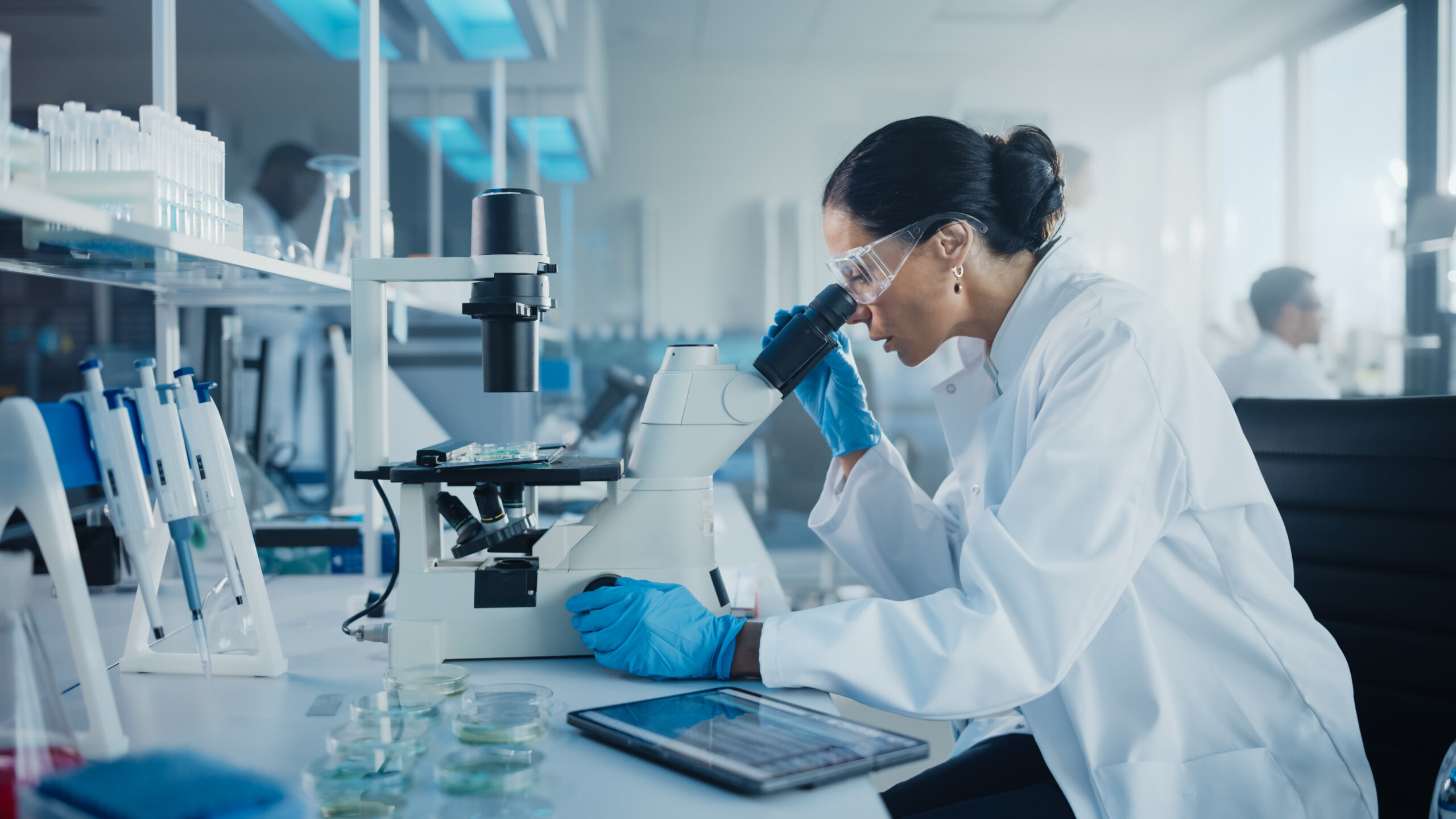 Medical Development Laboratory: Caucasian Female Scientist Looking Under Microscope, Analyzes Petri Dish Sample. Specialists Working on Medicine, Biotechnology Research in Advanced Pharma Lab.