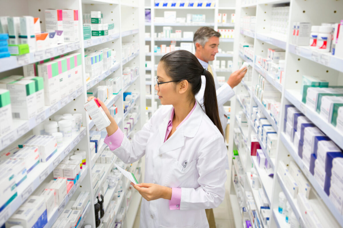 Pharmacist with a prescription looking at a medicine box on a pharmacy shelf.