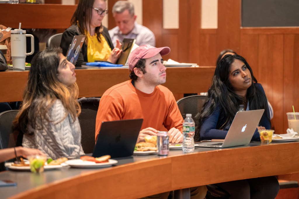 Students seated in a tiered classroom with laptops and lunch plates look up attentively toward the front of the room.