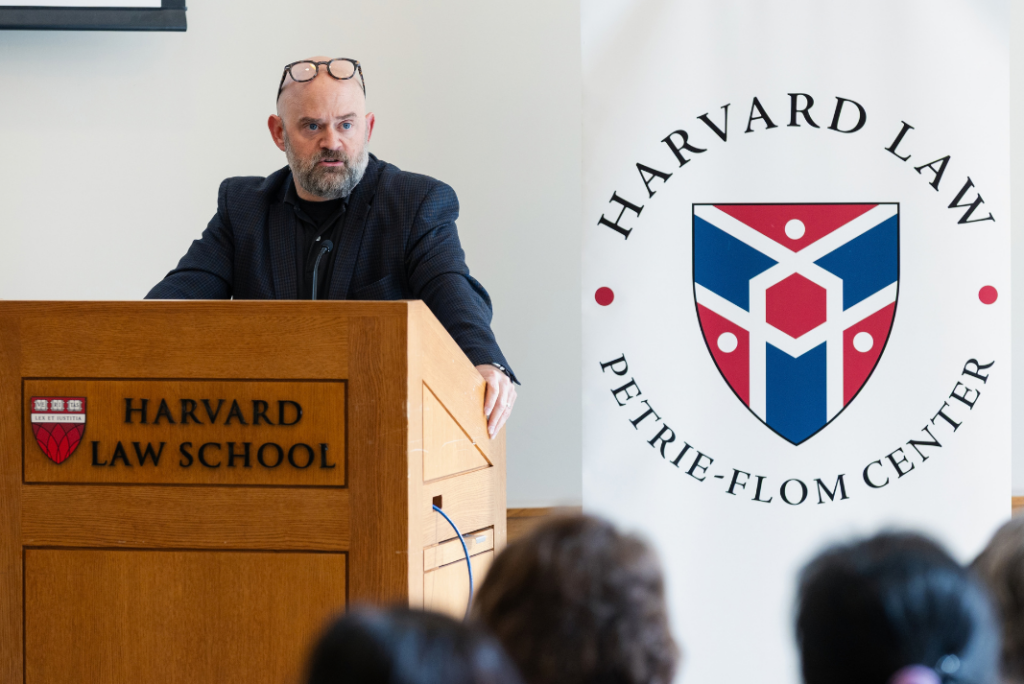 A bearded speaker stands at a wooden Harvard Law School podium beside a Petrie-Flom Center banner, with audience members visible in the foreground.
