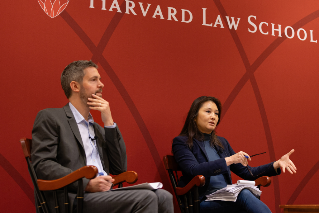 Two panelists sit in wooden chairs in front of a red Harvard Law School backdrop. A man in a gray suit rests his chin on his hand and listens while a woman in a navy blazer gestures as she speaks, holding notes in her lap.