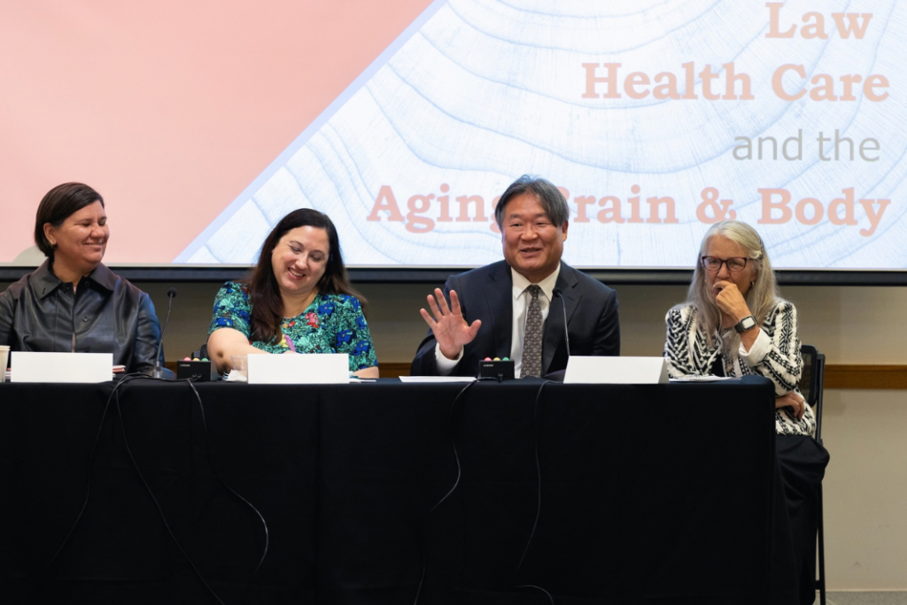 Four panelists sit at a table beneath a screen showing the title “Law, Health Care, and the Aging Brain & Body.” A man in the center raises his hand as he speaks while the others listen.
