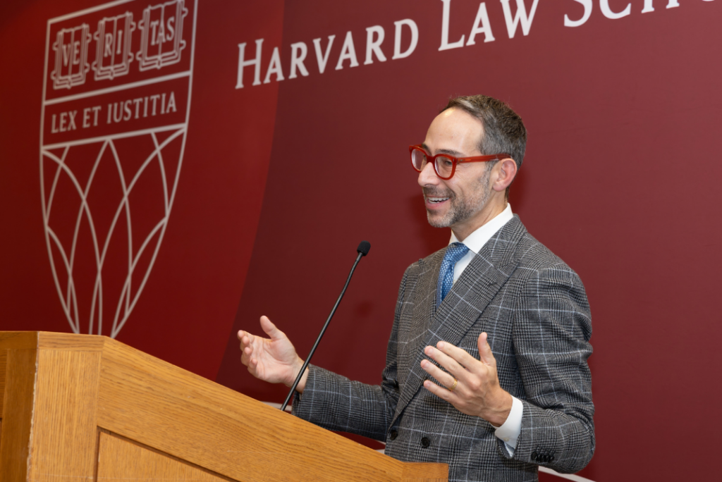 A man in red glasses and a gray plaid suit smiles and gestures as he speaks at a podium in front of a Harvard Law School backdrop with the school’s shield.