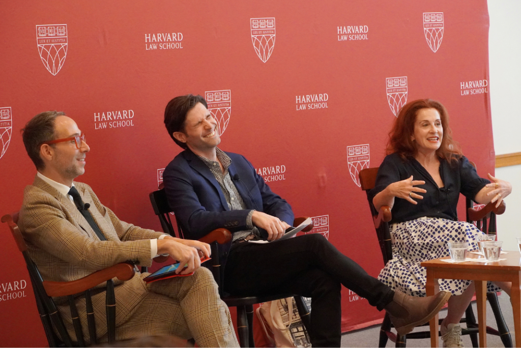 Three panelists sit in wooden chairs in front of a red Harvard Law School backdrop. The man in the middle laughs while the woman on the right speaks and gestures with both hands.