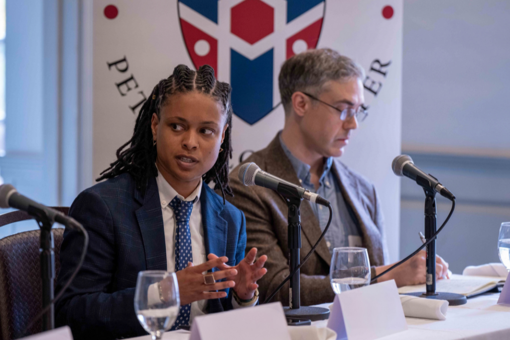 A panelist in a navy suit and polka-dot tie speaks and gestures at a table with microphones, while a man in a brown blazer sits beside her in front of a Petrie-Flom Center banner.