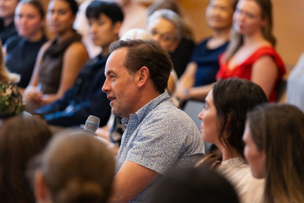 An audience member holding a microphone asks a question from his seat in a crowded room of attendees.