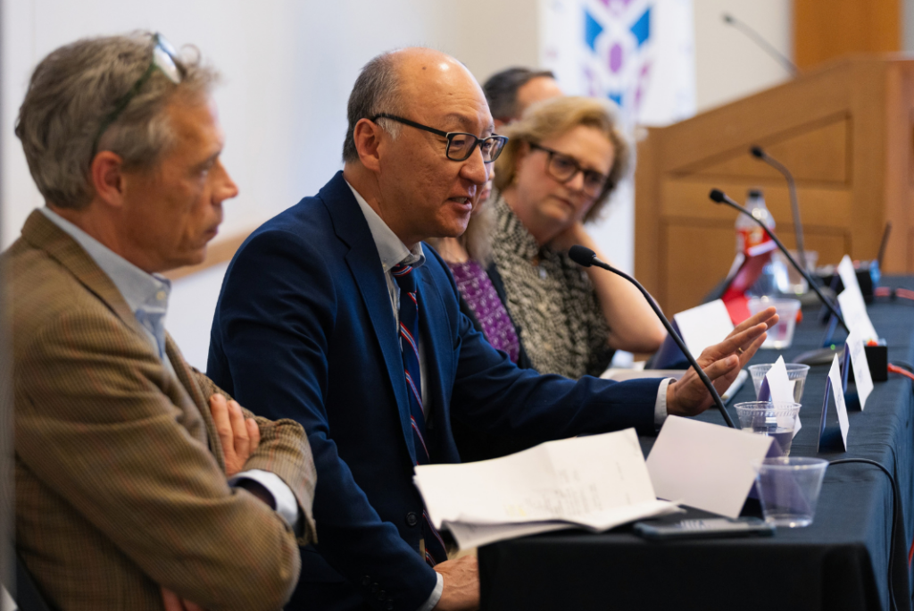 A man in a navy suit and glasses gestures while speaking into a microphone at a panel table, with fellow panelists seated on either side of him.