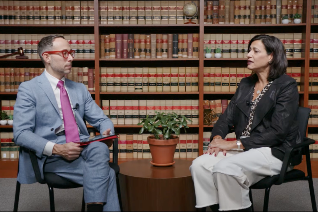 Two people sit facing each other in conversation in front of floor-to-ceiling shelves of law books. A man in a light blue suit and red glasses holds a tablet as a woman in a black leather jacket speaks.