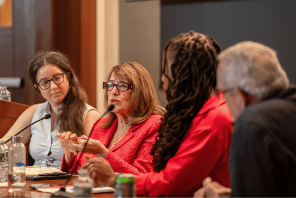 A woman in a red jacket and glasses speaks into a microphone during a panel discussion, while a woman in glasses beside her listens and two other panelists sit in the foreground.